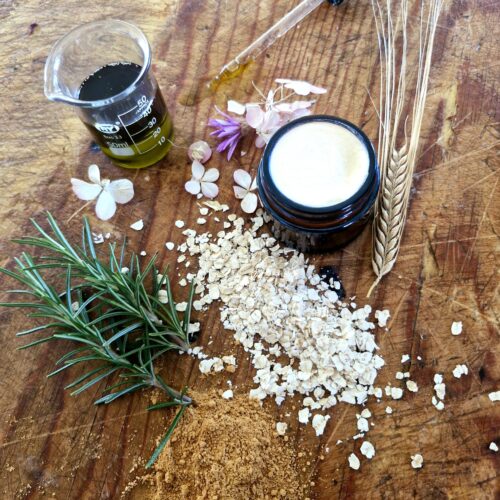 Flat lay of natural skincare ingredients including botanical oils, rosemary, oats, clay and an amber jar of grass-fed tallow cream on a wooden surface - Fatglow handcrafted skincare New Zealand